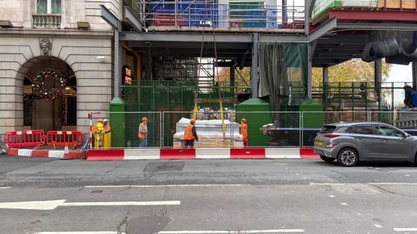 Building under construction with wire fence hoarding and several workers managing a load from a crane lift.